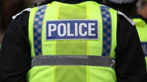 A stock image of a police officer seen from the back. He is wearing a high visibility vest with the word 'police' at the back, over a black jacket.