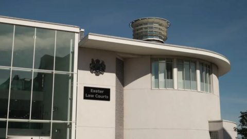 Exterior of the Exeter Law Courts building, featuring large glass windows on the left and a cylindrical structure with horizontal slats on top. A sign reading 'Exeter Law Courts' is visible below an official emblem.
