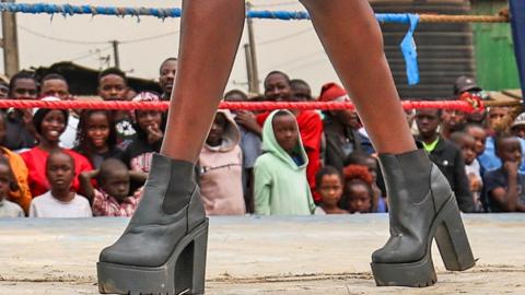 A close-up of a model's black heeled boots as she parades in a boxing ring watched by crowds of people in Nairobi, Kenya.
