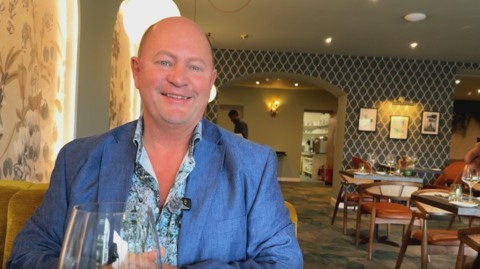 A bald man sat in a hotel restaurant smiling at the camera. He is wearing a blue patterned shirt and a blue suit jacket. 