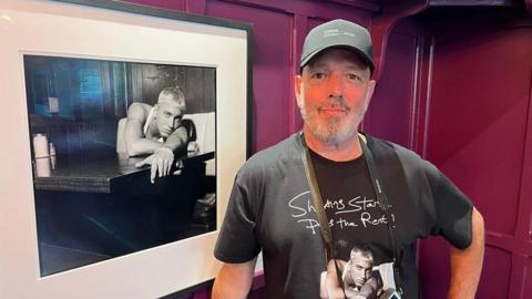 Steve Read stands beside a framed black-and-white photograph. Read is wearing a dark baseball cap and a black T-shirt featuring artwork from the exhibition. The photograph beside him shows a blond-haired musician leaning on a counter, while burgundy-coloured wall panels form the background.