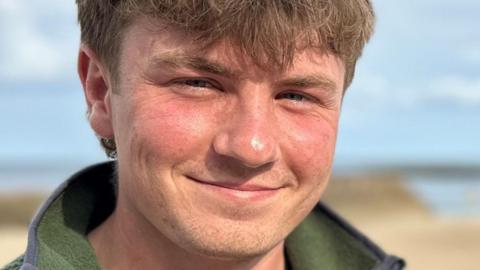 Euan and Andrew stand side by side smiling at the camera, with a yellow beach and blue sea behind them. Euan has brown hair and is wearing a green fleece. Andrew has black hair and is wearing a grey hoody beneath a navy body warmer.