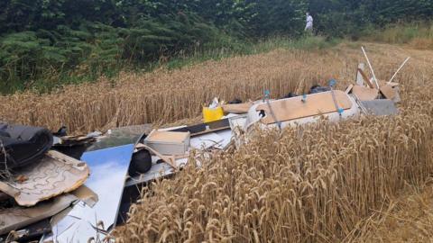 A pile of rubbish including a bath and pieces of wood on the ground in a field. There are trees in the background. 