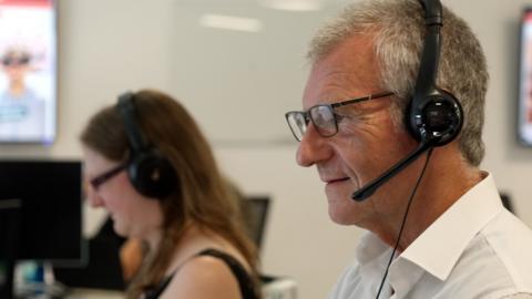 two call handlers can be seen, the man in the foreground is wearing a headset, a white shirt and has grey hair, a blurred woman in the background is wearing a headset and a black top