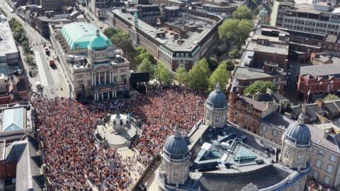 A huge crowd of people fills a city square around a central monument and historic buildings, with a grand domed building and surrounding streets packed tightly in a large public gathering.