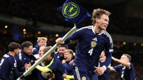 Scott McTominay celebrates with the corner flag. Other Scotland's men's national team players celebrate in the background. They are all wearing navy blue Scotland football shirts and shorts with three white stripes.