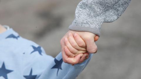 A stock image of a child in a blue jumper with stars on it holding the hand of an adult in a grey jumper. It is zoomed in on their hands and their faces cannot be seen. The background is out-of-focus and grey.