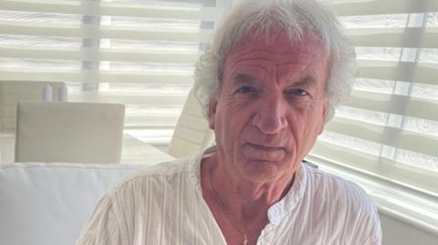 Image shows Stephen Copson. he is 72 and has white hair, wearing a white shirt, sitting in his house in Thornton. 