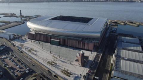 An aerial view of Everton Stadium, a curved silver structure on dockland with the River Mersey in the backdrop