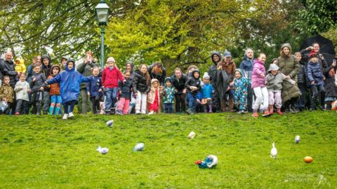 A group of people wearing coats and winter clothing stand at the top of a grassy hill, with large, designed eggs rolling down it. Some of the eggs are in plastic bags. There are leafy trees and a lamppost in the background.