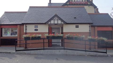 The public toilet is a red-bricked building with a tiled roof and a cream concrete exterior décor. A red door in the middle opens up onto the toilets. There are planters on top of the black metal fence out front.