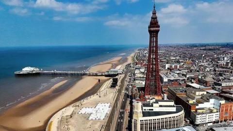 A general aerial view of Blackpool showing the tower and the town to the right, and the beach to the left with central pier. The sky is blue with a few white clouds.