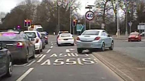 A silver Vauxhall Astra is mounting the central reservation on a busy multi lane A-road. The image is blurry as it has been captured by a police dash cam. 