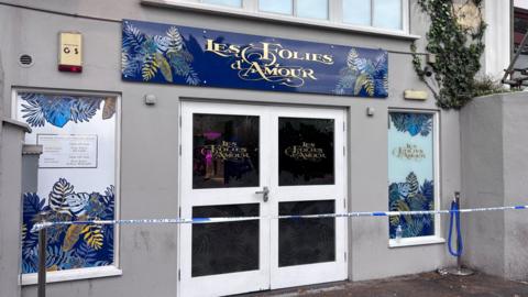 Police tape outside Folies nightclub in Guernsey. The building has double doors with white frames. It has a blue sign with the club's name on with gold writing. It also has floral patterned designs in windows either side of the doors.