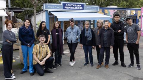 Eleven people who work in independent businesses at Gabriel's Wharf pose in a line and look at the camera. They stand in front of a shop with the sign: Southbank Printmakers.