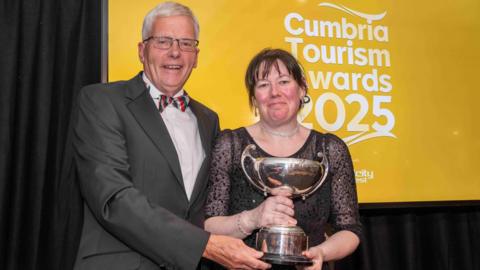 Jim Walker (left) and Tracy Hodgson holding a silver trophy. Mr Walker has grey hair and glasses and is wearing a grey suit, white shirt and striped bow tie. Ms Hodgson has brown hair and is wearing a black dress. They are standing in front of a sign reading "Cumbria Tourism Awards 2025".