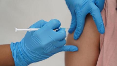 A close up of someone being given an injection. The person giving the injection has blue gloves on and the person receiving the injection is wearing a pink top. 
