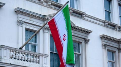 A close-up shot of the upper floors of the Iranian Embassy focusing on an Iranian flag hanging from a pole that sticks out from a balcony