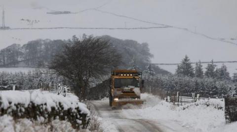 A gritter ploughs through snow on a rural road.