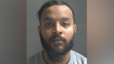 A custody image of Mustak Alom. He wears a grey sweater and has a beard and moustache.