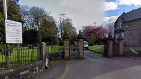 The entrance to a cemetery, with gates and a building in front of grass and trees in the distance 