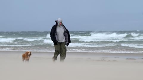 Man and his dog walking along a beach where sand is visibily lifted up with large waves in the background