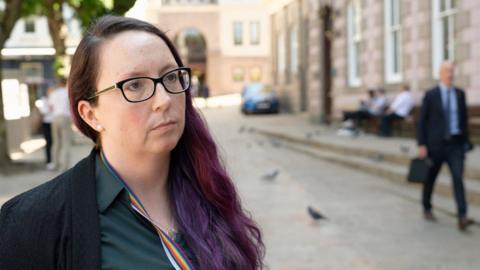 Deputy Doublet has glasses and a green shirt. She is stood in Jersey's Royal Square. 