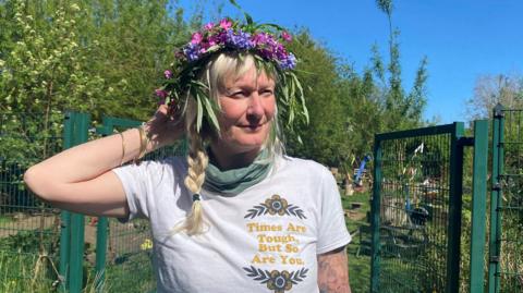 Cath Darling is a woman in her 50s with blonde hair in a plait and wearing a white t-shirt with the words Times are tough but so are you on it. She is standing in a garden with a garland of flowers on her head.
