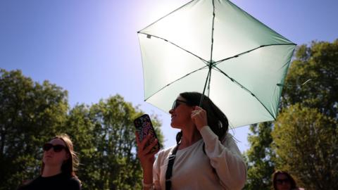 Two women wearing sunglasses on a sunny day. One carrying an umbrella and mobile phone