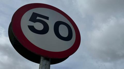 Low-angle view of a circular 50 mph speed limit sign against a cloudy sky, with a tree visible to the side.