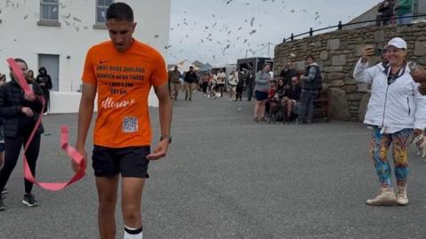 Amol Randhawa crossing the finish line in Land's End, Cornwall. Amol is wearing black shorts and an orange t-shirt with Ellenor emblazoned on the front. A red ribbon and confetti can be seen in the picture. 