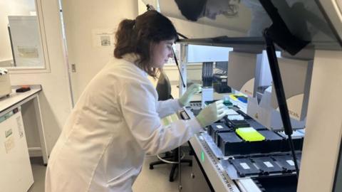 A woman in a laboratory bending over a machine holding test tubes and other laboratory equipment. She has long dark hair, and is wearing a white lab coat and protective gloves.