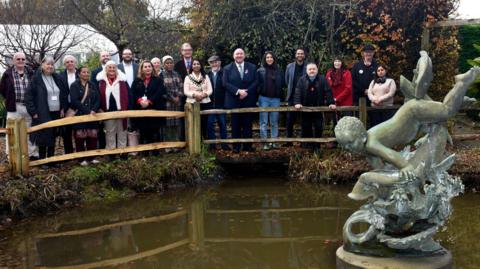 The metal statue of a boy jumping over the top of a fish sits in the middle of a park pond. A large group of people, some wearing chains of office, are lined up along a bridge over the water feature, looking straight at the camera.