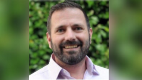 Martin Realey smiles for the camera for an official headshot photo. He has short trimmed black hair, and a neatly trimmed black and grey beard. He is wearing a pink smart shirt. 