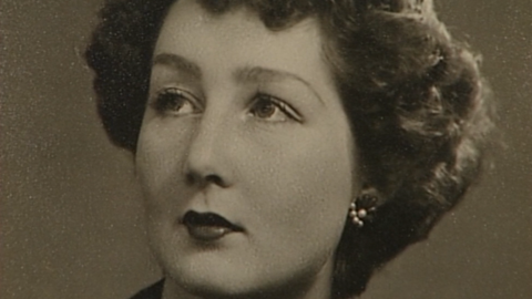 A young Noreen Riols gazes upwards and sideways in a pose in a brown-tinged monochrome photograph. She wears lipstick and other make-up, has earrings and her wavy, brown hair is cut in a short style.