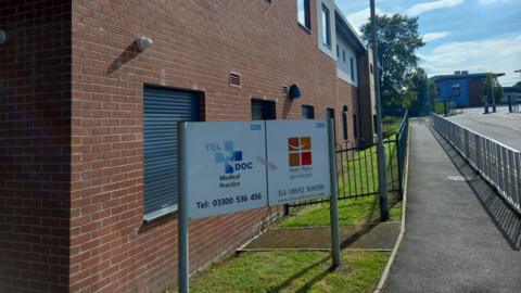 The sign outside Teldoc Medical Practice, alongside a pavement separated by railings from a road