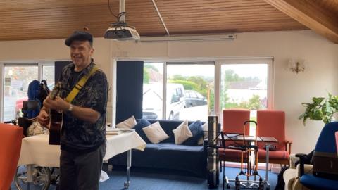 Performer playing an acoustic guitar in a lounge-style room with armchairs, a sofa and large windows, with a music stand and walking aids visible nearby.
