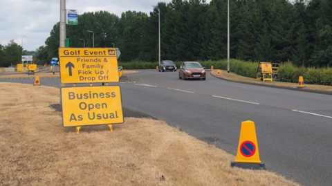 Two yellow signs are by a road near a mainly yellow traffic cone. One sign includes the words golf event and the other includes the words business as usual. A roundabout and trees are in the background.