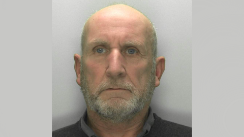 A police mugshot of Michael Florek. He has a bald head with grey hair on the sides, and a grey beard. He is wearing a dark collared shirt and standing against a grey background with a blank expression.