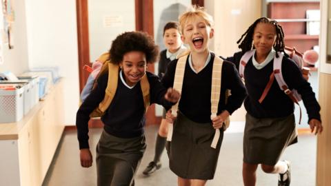 Children run down a school corridor with their backpacks on. All of them look excited and are laughing.