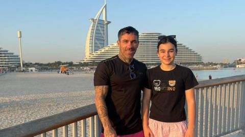 Ryan Hodgson stands next to his daughter in front of The Palm Dubai. They are both wearing black T-shirts. There is a beach behind them and blue skies.