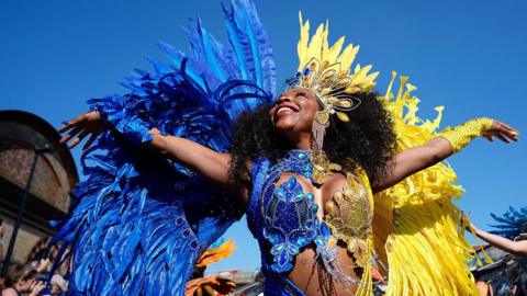A woman in a blue and yellow feathered carnival outfit, head looking up to the sky and smiling, arms outstretched.