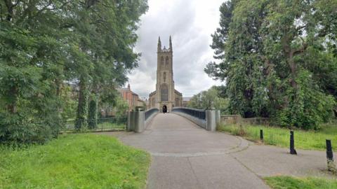 A Google maps photo of a footbridge. Before the bridge is grass and trees either side. At the far end of a the bridge, there is a church