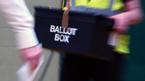 A stock image of a ballot box arriving during the count, carried by a person wearing a high-viz vest.
