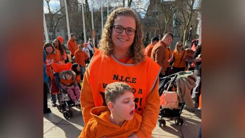 A woman in a orange jumper and orange vest holds a small boy in a orange hoodie. Behind them are more people dressed in orange clothing. 