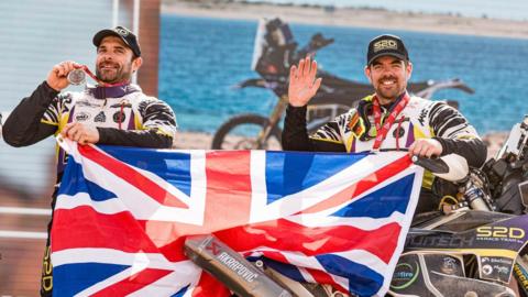 Two men in motocycle gear and caps both have medals and are holding up the Union Jack flag