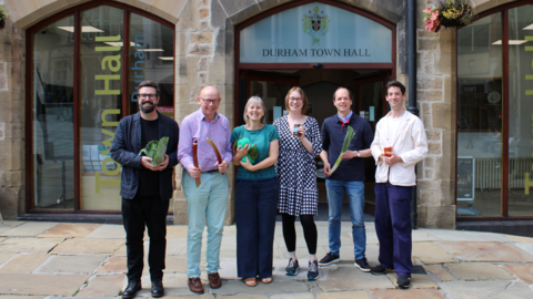Organisers Rob King, David Ridgeway, Nicola Tomaney, Helen Billam, Barend van Leeuwen, Benedict Douglas smiling outside Durham Town Hall. They are each holding a vegetable or a jar of jam. Rob King has black hair and beard, glasses and wears an indigo jacket over a black top and trousers. David Ridgeway, has glasses red and blue checkered shirt and light blue trousers. Nicola Tomaney's straight grey hair reaches her shoulders and she wears a blue top and wide leg indigo jeans. Helen Billam has glasses, short brown hair and wears a black and white checkered dress over black leggings. Barend van Leeuwen has short brown hair and wears jeans and a blue long-sleeved top over a light blue shirt. Benedict Douglas has black hair. He wears wide leg indigo jeans and a cream cardigan over a white top.