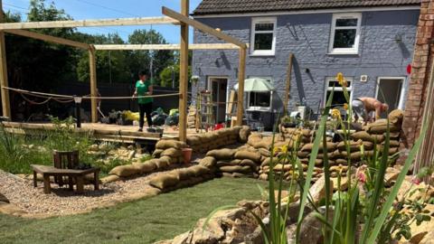 A garden with sandbag lined paths and walls. There is a pergola on a raised area and plants. 