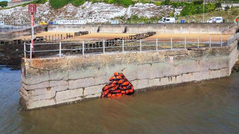 A sea wall in Ramsgate with a pile of lifejackets painted on it in orange and black. The painting is in a 3D effect and the beach is seen in the background along with low cliff faces