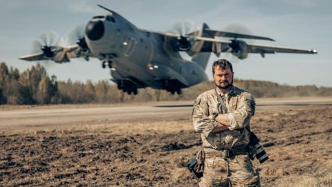 A man, Aaron Stone, stood with his armed crossed with two cameras hanging from his side. A large cargo plane is taking off in the background.